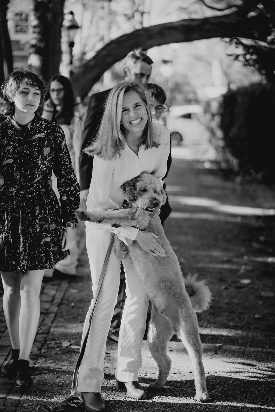 Black-and-white image of a mom hugging her dog with the family behind her, capturing a candid moment full of joy.