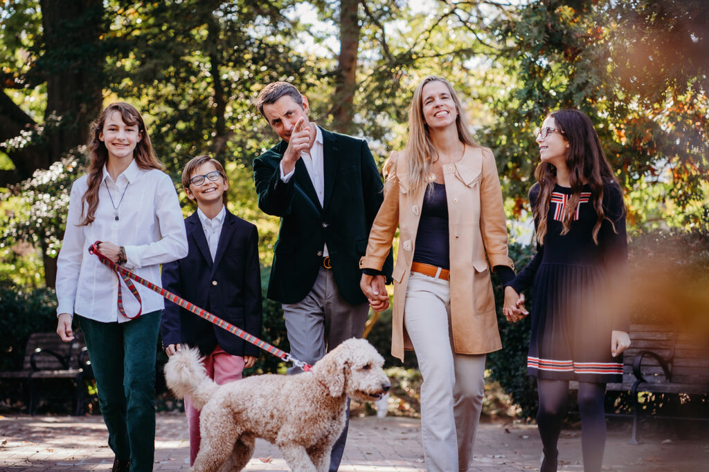 Family strolls hand in hand with their dog on a shaded park path, laughing and enjoying a relaxed moment during their annual family photos.