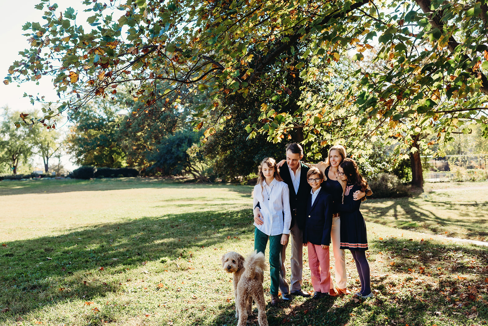 Family stands together in a sunlit field beneath a large tree, smiling for their annual family photos with their beloved dog in front.
