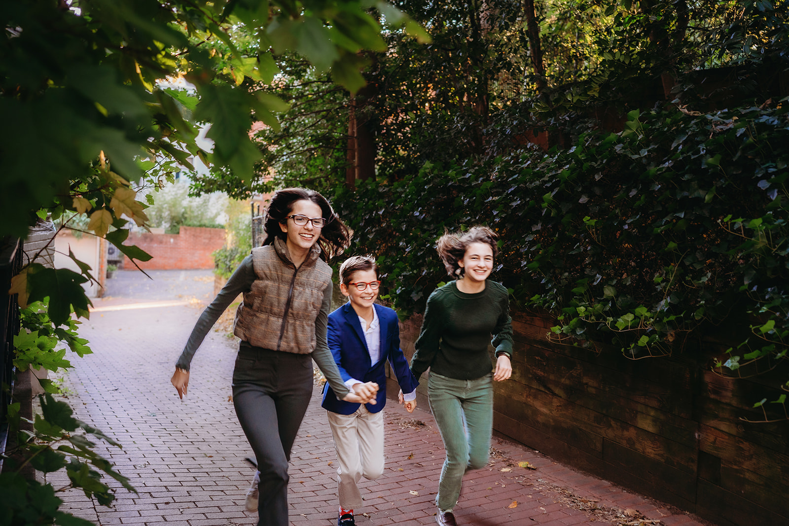 Three siblings run through a brick alley holding hands, caught in a playful moment during their annual family photos.