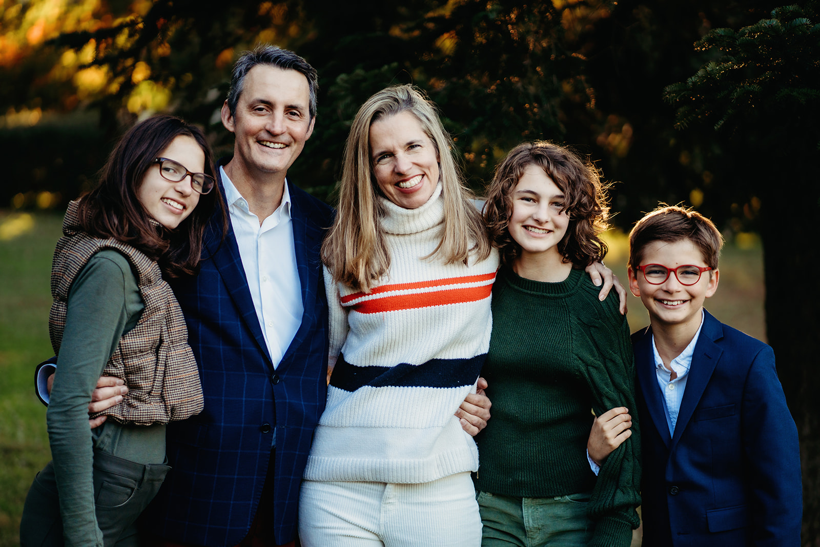 Parents and three kids pose together with big smiles, glowing in the soft golden-hour light near a wooded area.