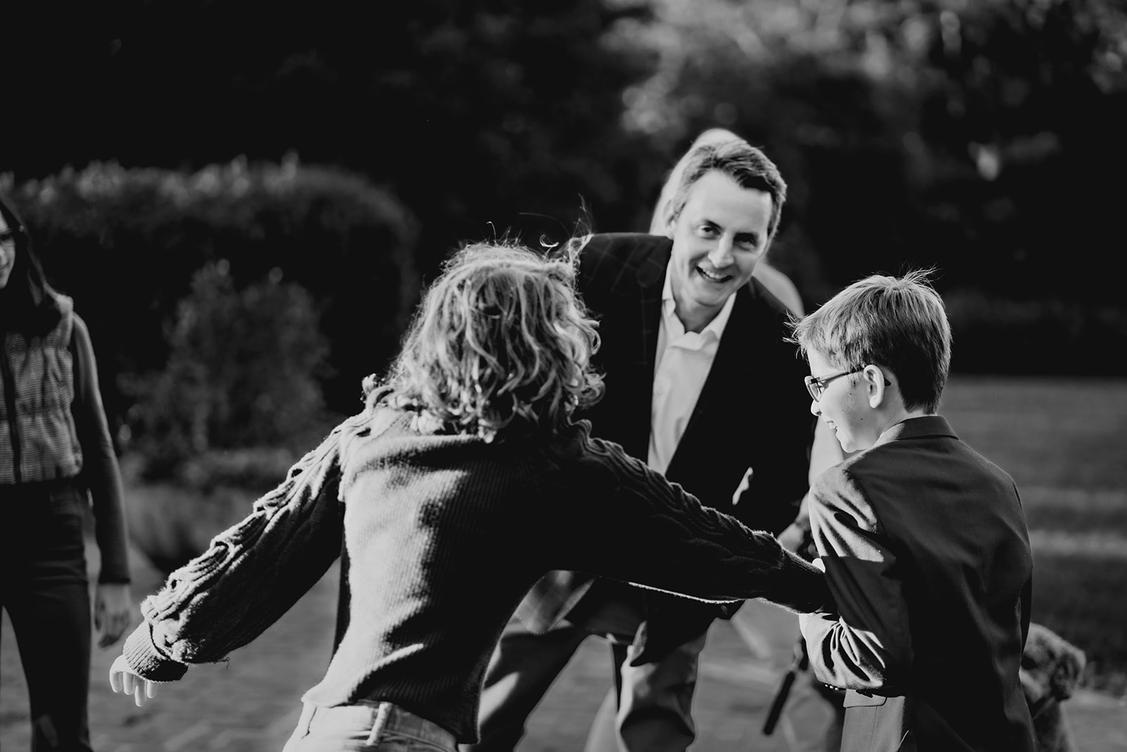 Black and white image of dad smiling as two kids playfully run toward him, capturing a spontaneous moment of connection.