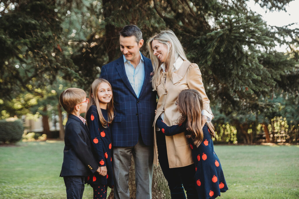 Parents and three kids share a warm moment under tall evergreens, dressed in fall layers for their annual family photo session.