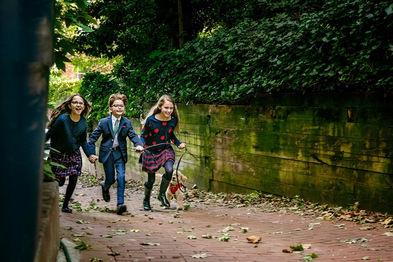 Siblings run hand-in-hand down a brick path with their puppy, a joyful moment from their annual family photos.