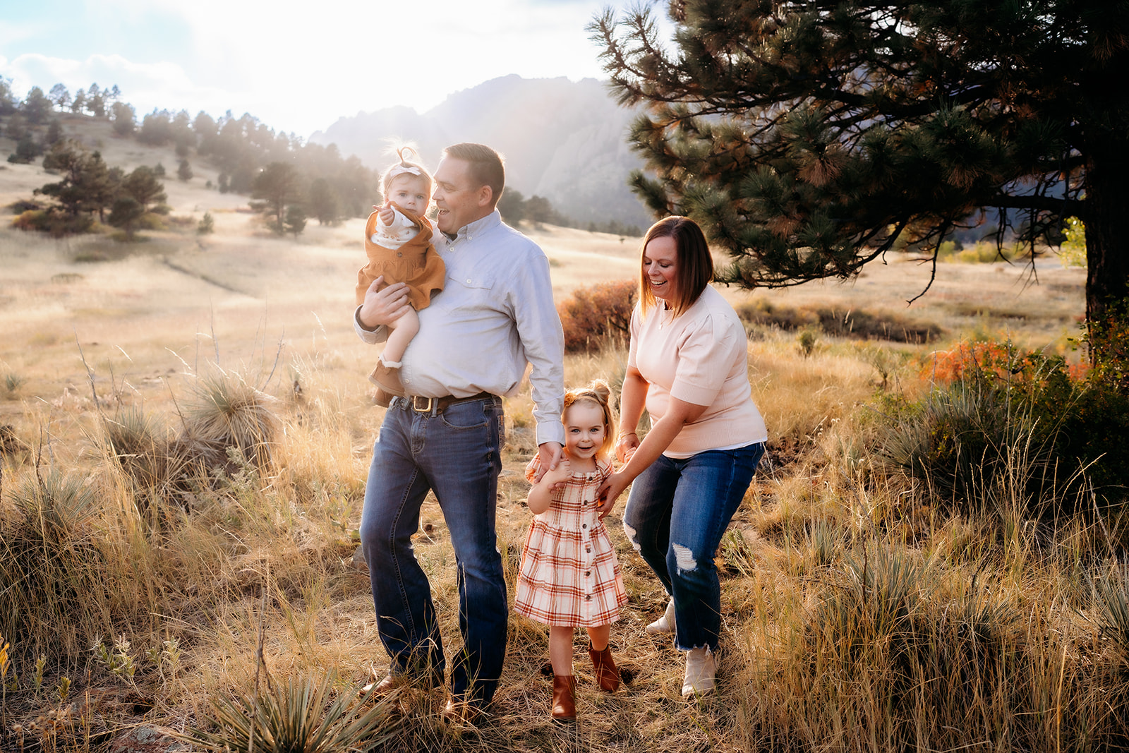 Family of four walking hand in hand through tall grass at golden hour with the Flatirons in the background during family photos Denver.