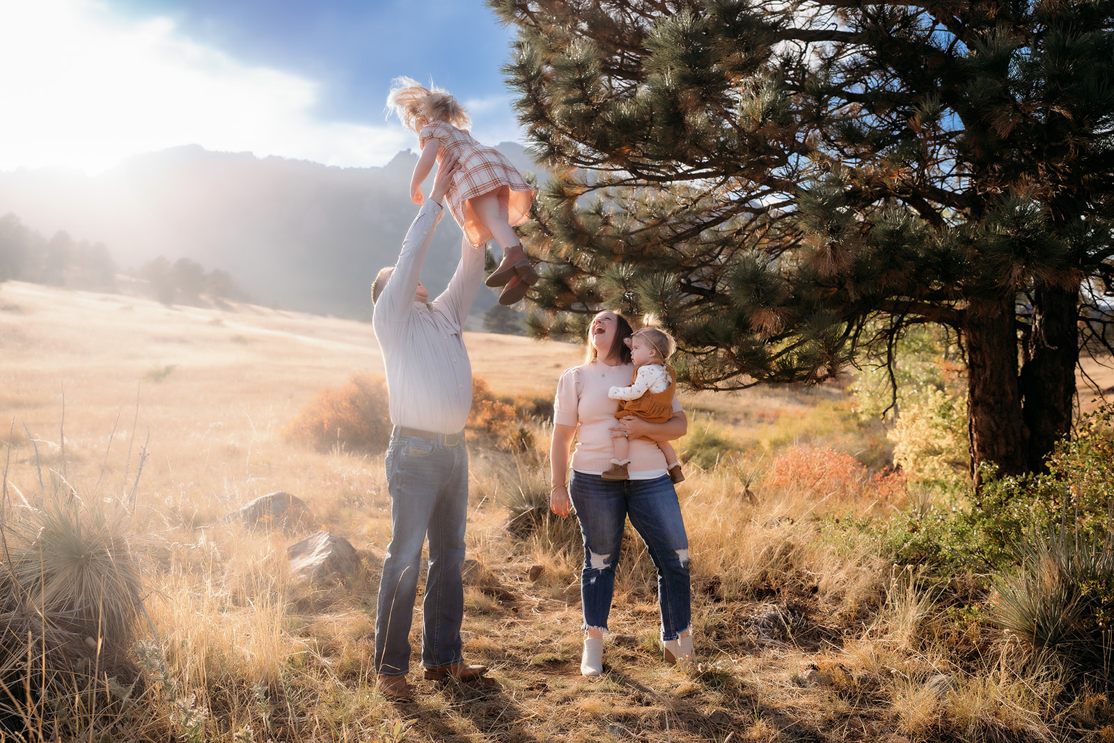 Dad tossing his daughter high into the air while mom holds their toddler beneath the trees during family photos Denver.