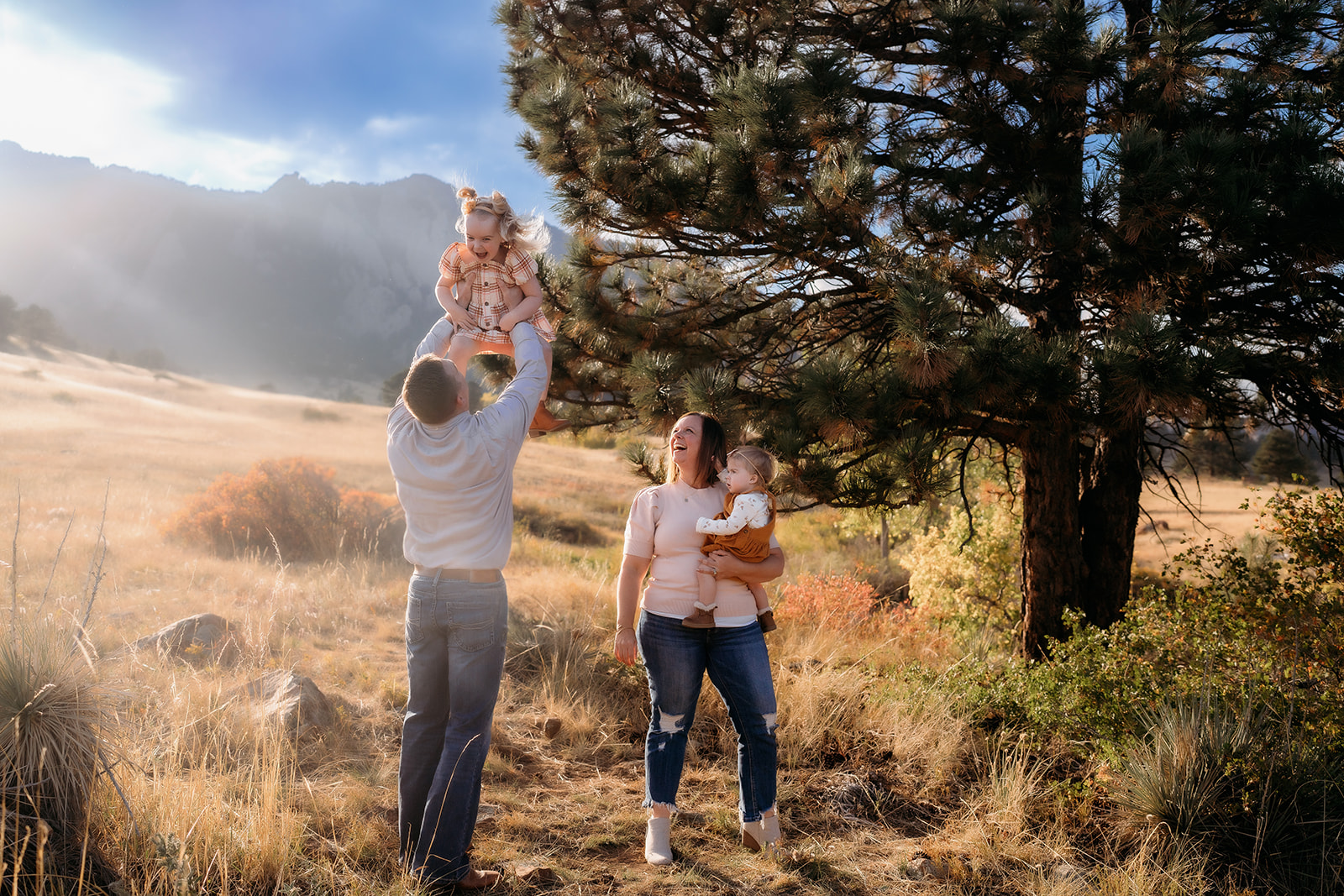 Dad lifting daughter high into the air while mom smiles and holds their toddler beneath the Flatirons.