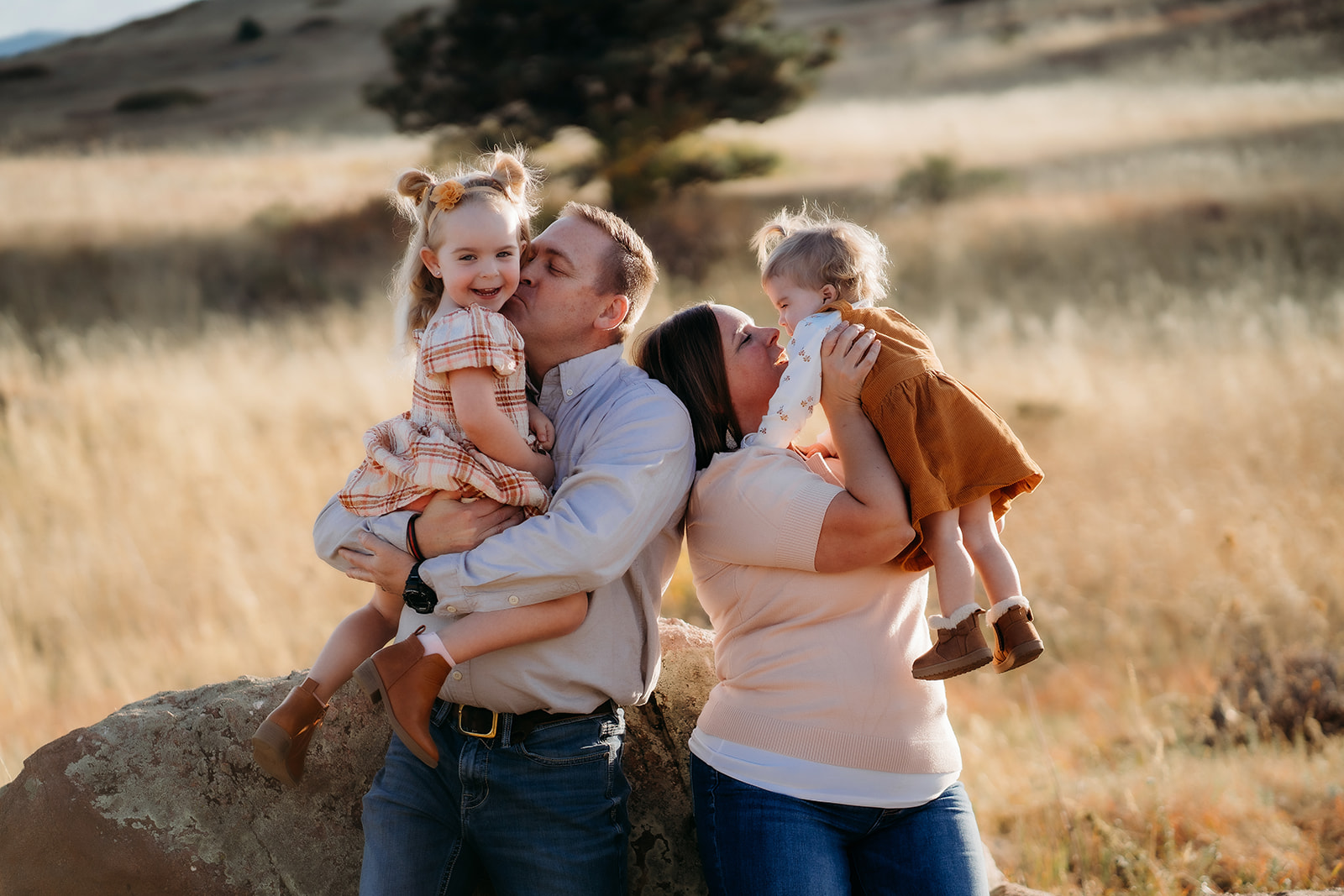 Parents kissing their daughters while standing near a boulder, surrounded by golden light during family photos Denver.