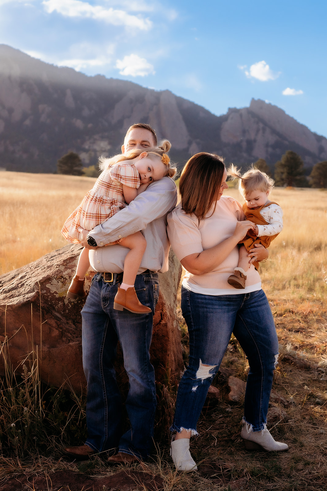 Parents holding their daughters close while standing near a boulder with Colorado’s Flatirons behind them.