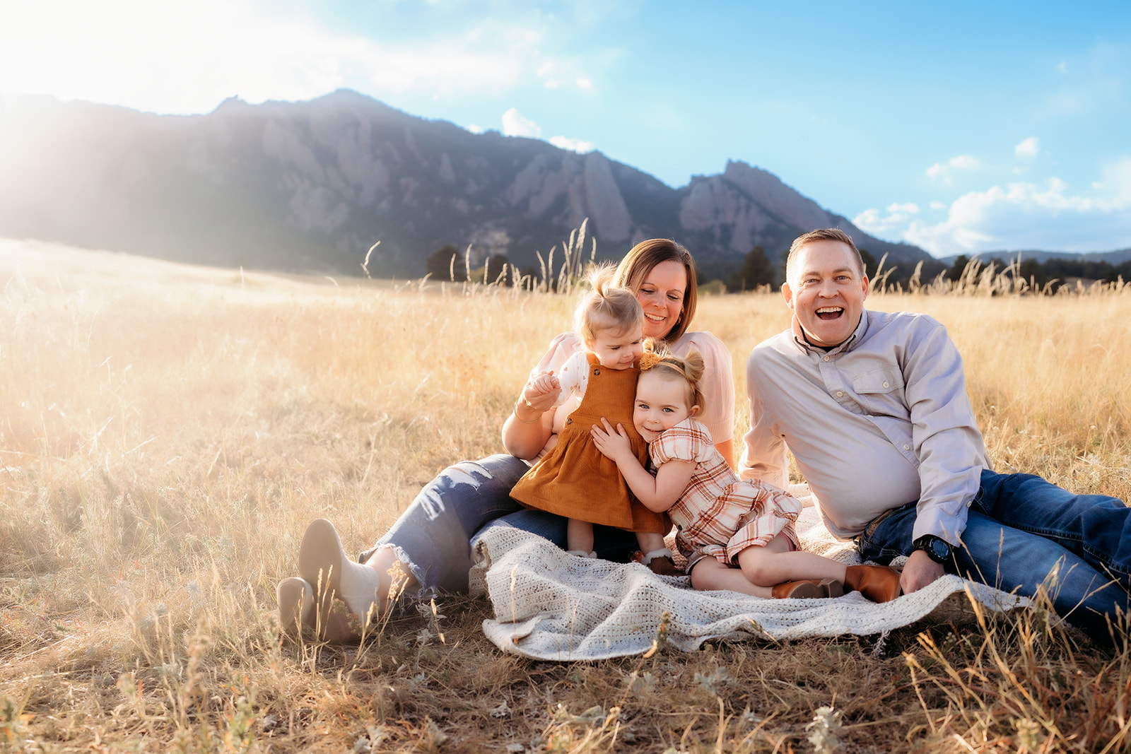 Parents and two daughters laughing together on a blanket in a golden field at the base of the Flatirons.