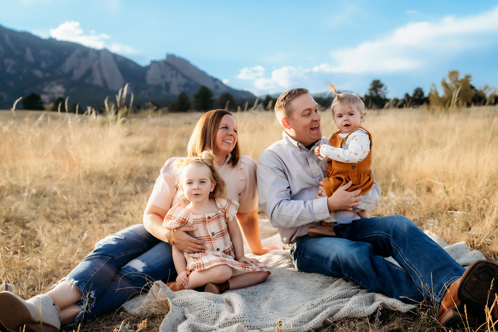 Family sitting together on a blanket in a golden Colorado field, laughing and holding their daughters.