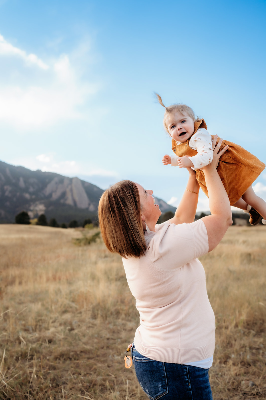 Mother holds her toddler high in the air against a mountain backdrop, both smiling in the autumn light during family photos Denver.
