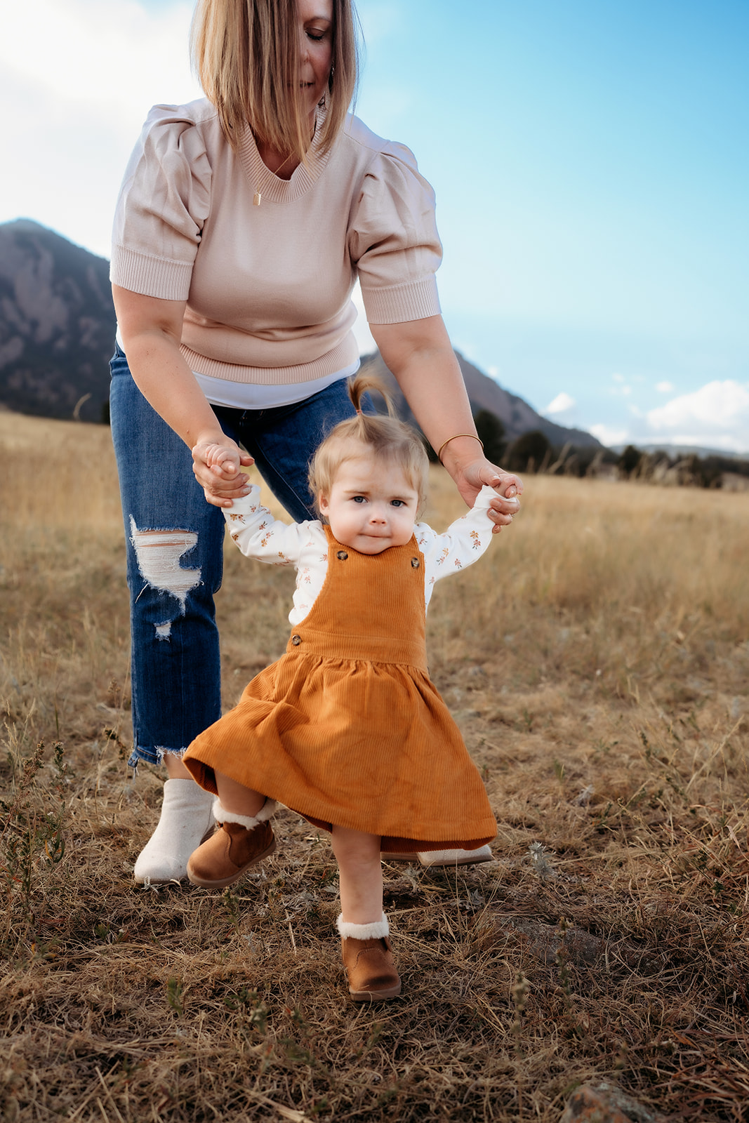 Mother holding her toddler’s hands as she takes confident steps in a fall meadow.