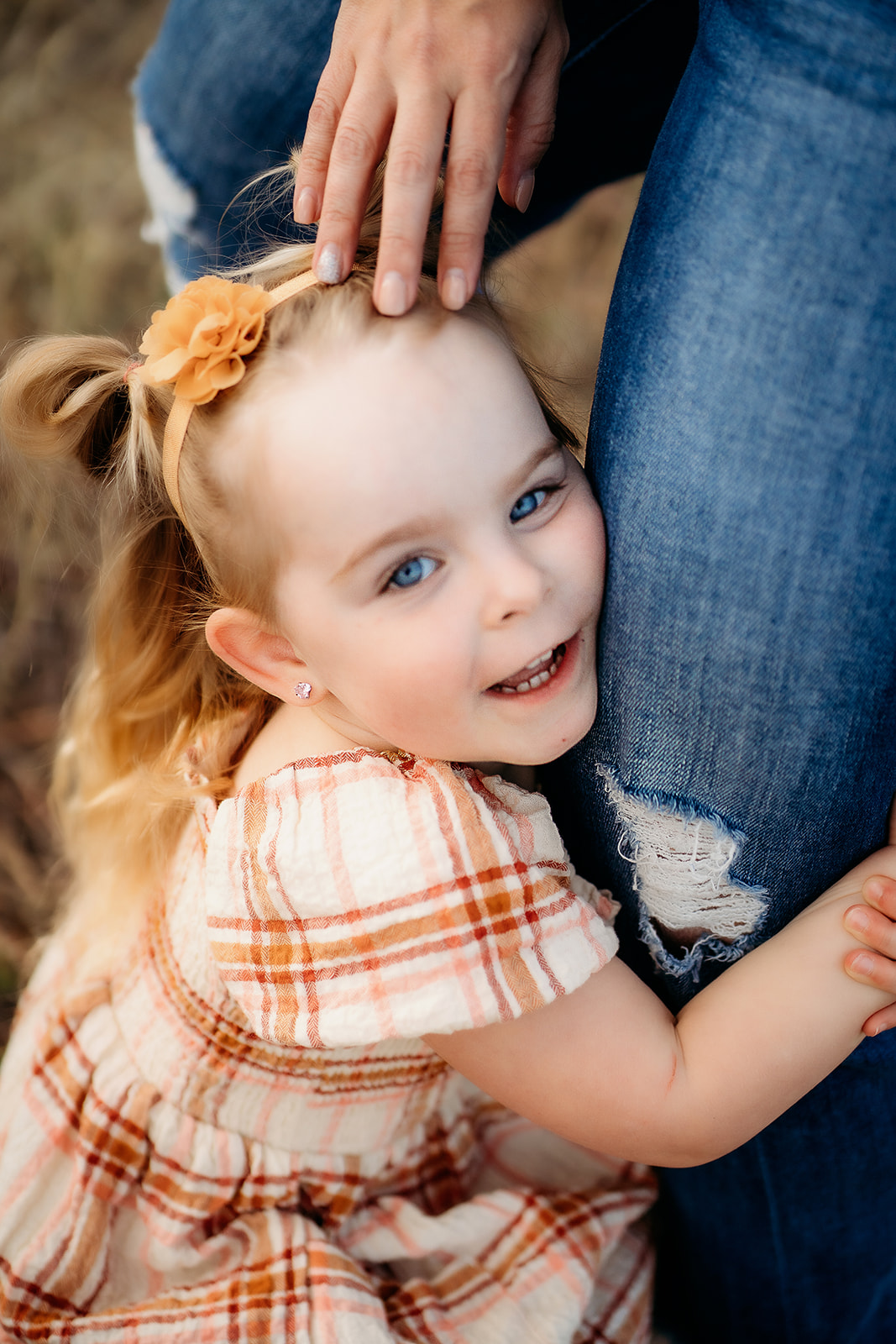Little girl in a plaid dress hugging her mother’s leg while smiling at the camera.