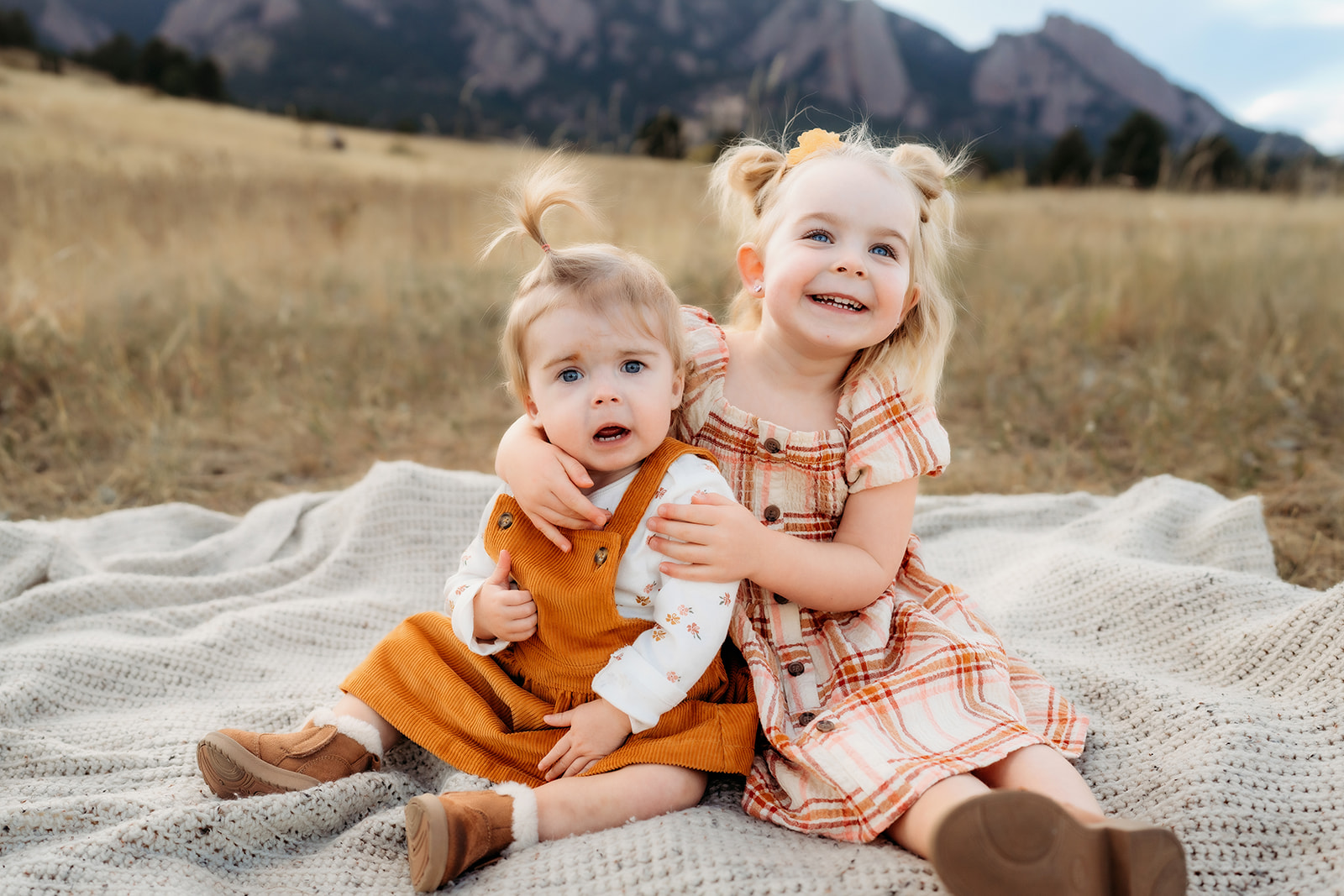 Two young sisters hugging on a blanket in the grass with Flatirons in the background during family photos Denver.