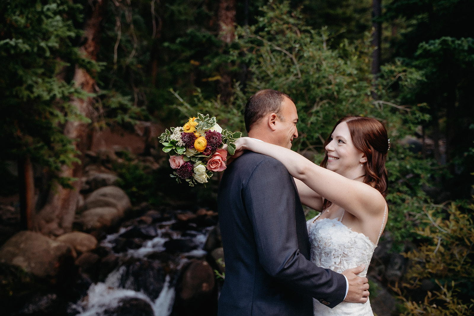 The bride wraps her arms around the groom’s shoulders, bouquet in hand, during their forest elopement in Colorado.