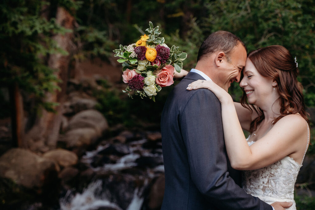 Her arms draped around his shoulders, bouquet raised in celebration, they smile ear to ear beside a bubbling creek.