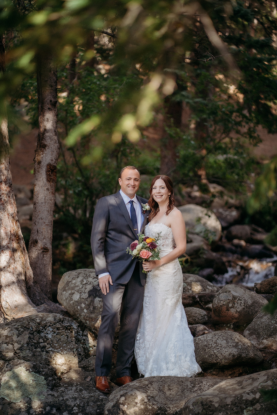 Framed by forest trees and creekside rocks, the bride and groom smile brightly for a peaceful wedding day portrait.