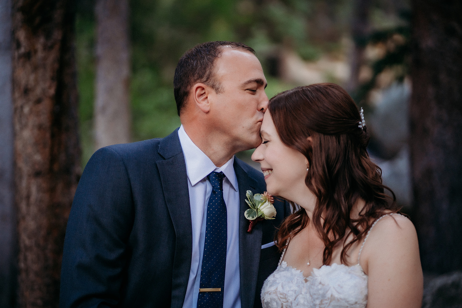 A tender kiss on the forehead, the groom shows his love as they embrace in the woods.