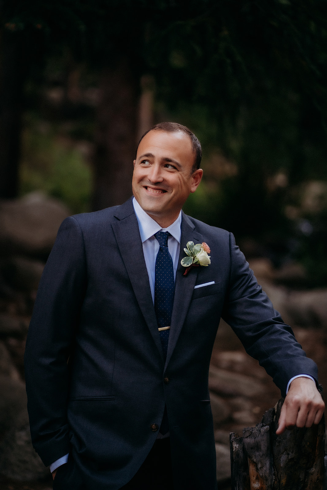 The groom leans casually against a log, smiling softly as he waits to see his bride.