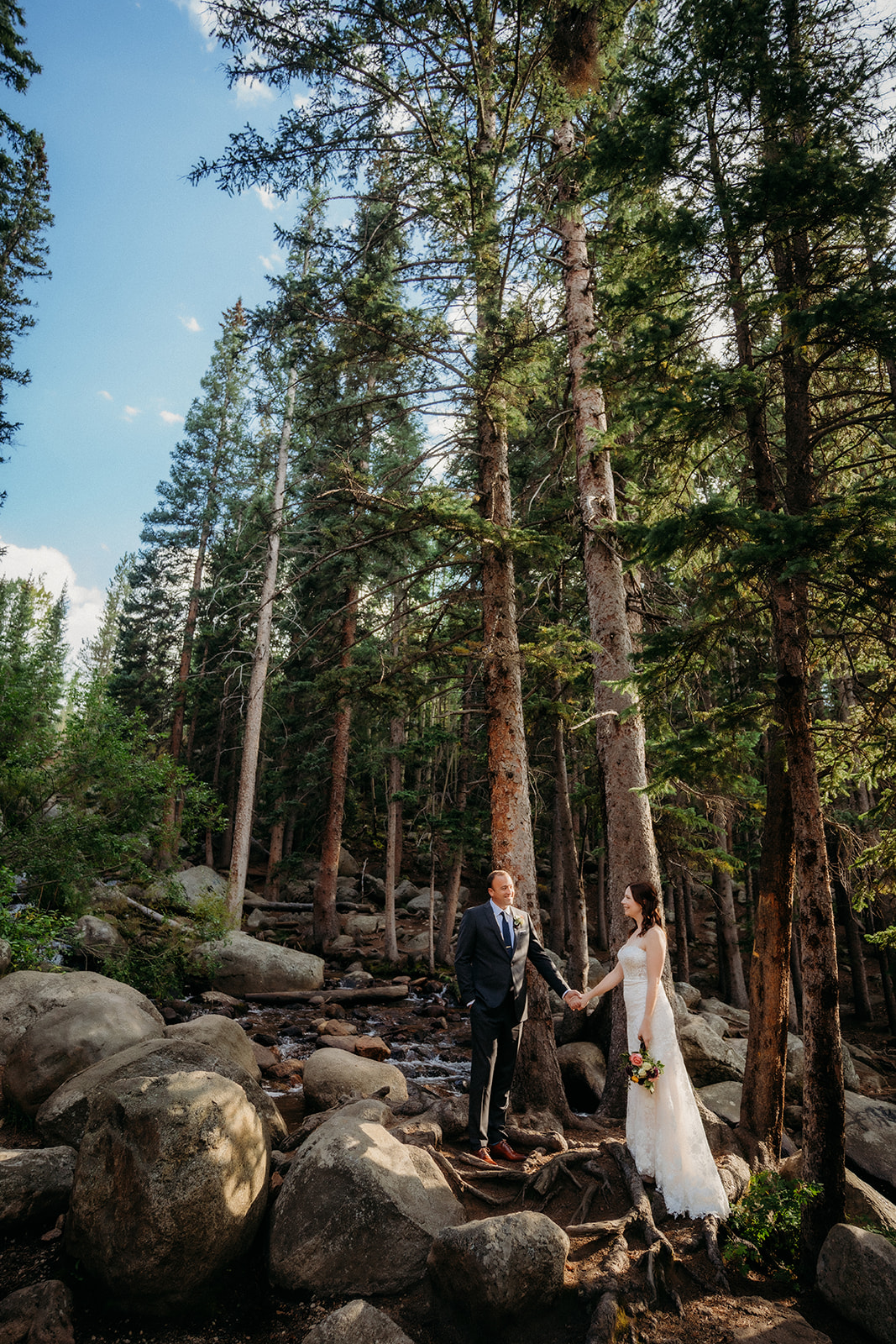 Standing on boulders beside a flowing creek, the couple holds hands in a moment of calm surrounded by nature.