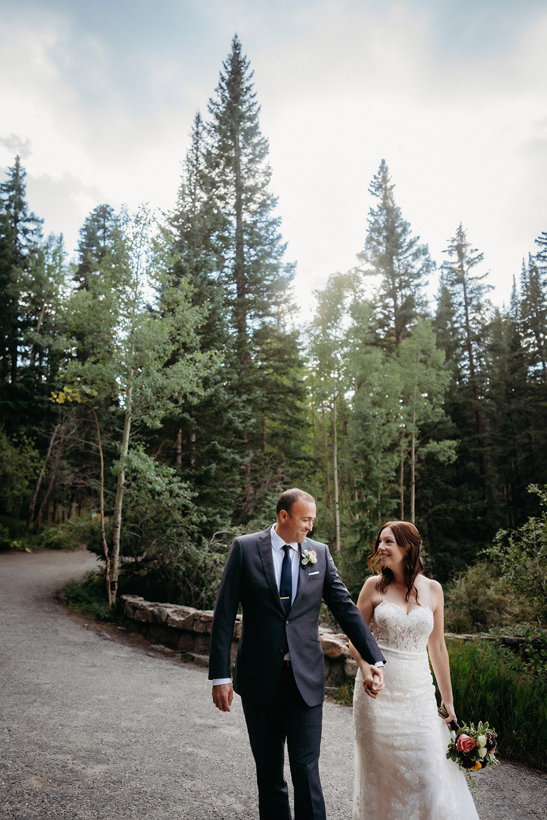 Walking side by side on a pine-lined trail, the newlyweds share smiles and sunlight.