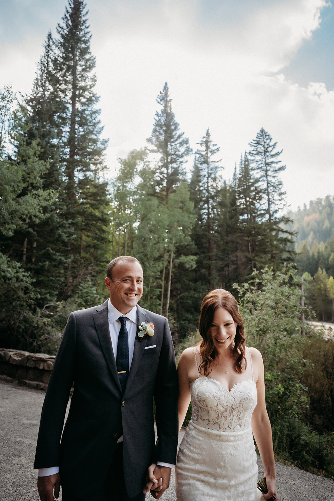 The bride and groom walk hand in hand through a quiet forest trail, surrounded by towering pine trees.