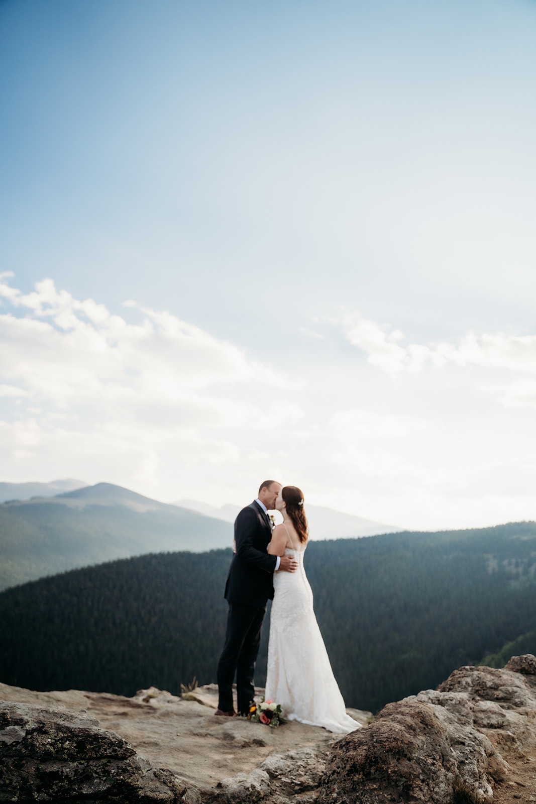 The couple shares a kiss on the edge of a cliff, surrounded by sky and rolling peaks—classic Colorado magic.