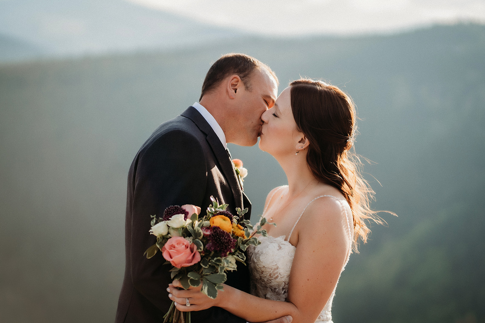 A romantic kiss with wildflower bouquet in hand, framed by hazy Colorado peaks.