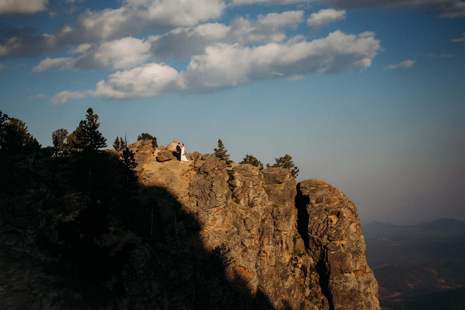 Perched on a distant cliff edge, the couple embraces as golden hour light hits the mountains around them.