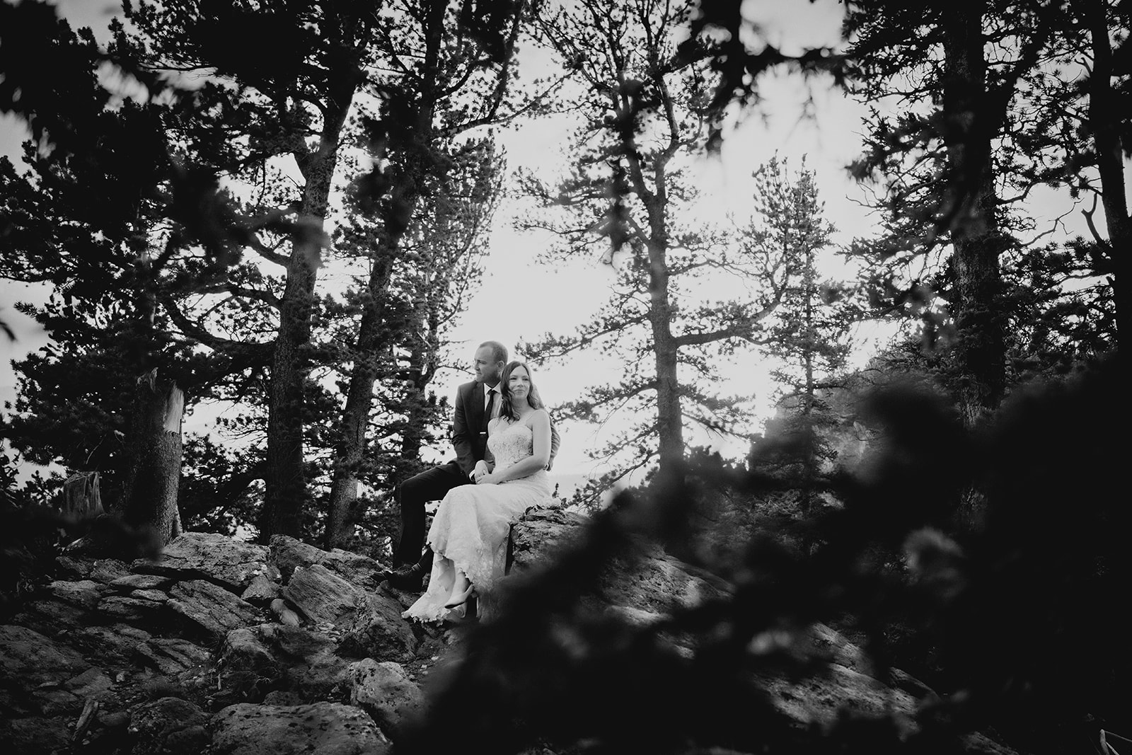 A quiet, black-and-white moment of the couple seated together on a rocky forest ridge.