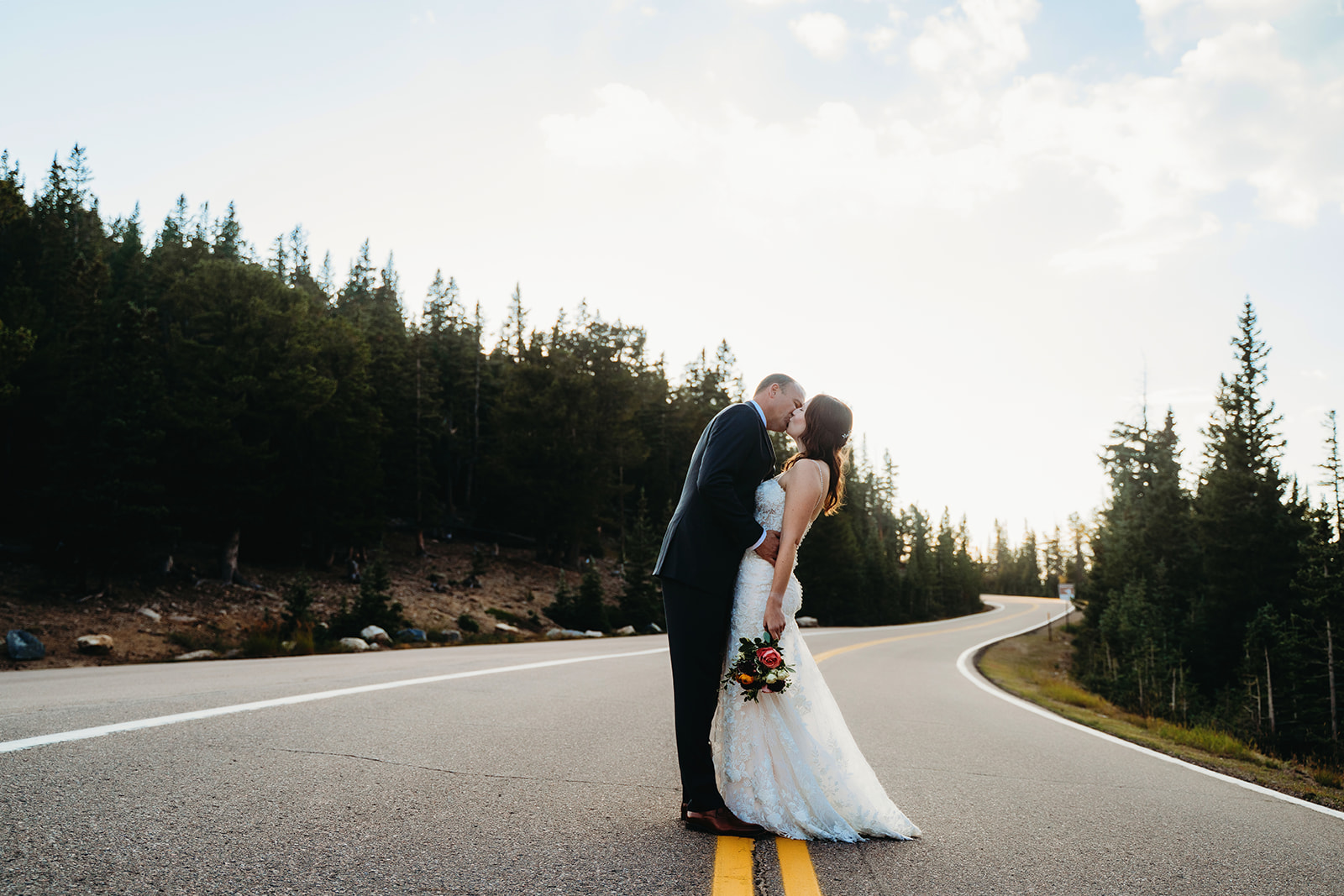A couple shares a kiss on an empty mountain road, the winding pavement and tall pines framing their moment.