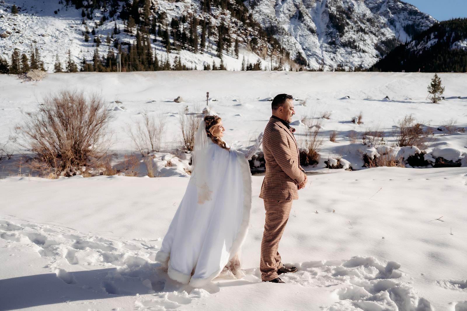 Bride stands behind the groom with excitement before a first look, snowy mountains stretching out behind them.
