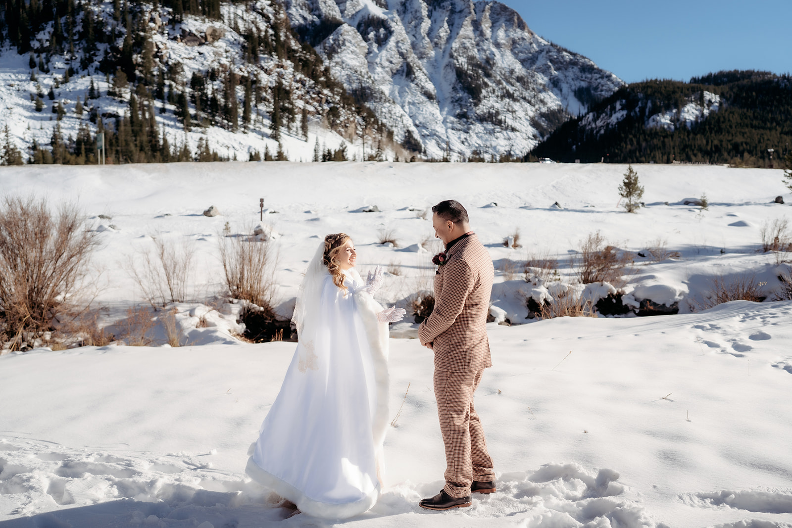 Bride and groom face each other in the snow, framed by dramatic mountain peaks and winter sunlight.