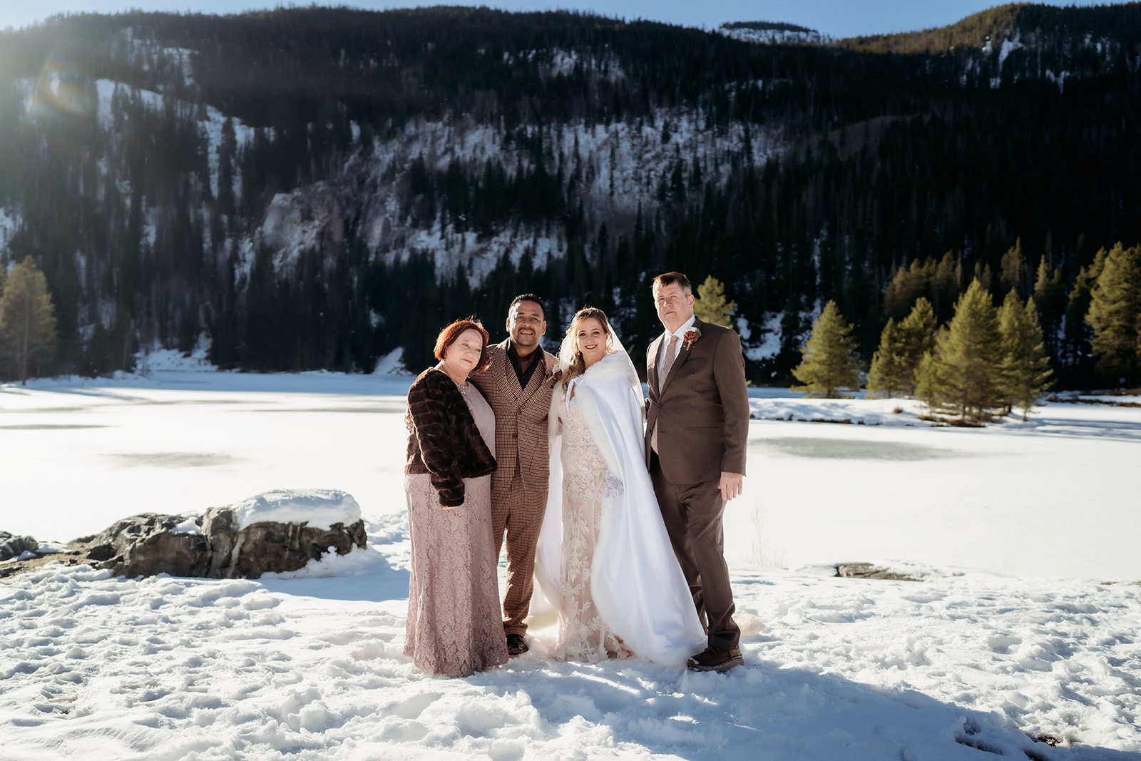 Bride and groom pose with two guests on a snowy lakeshore, surrounded by pine trees and winter sunshine in Colorado.