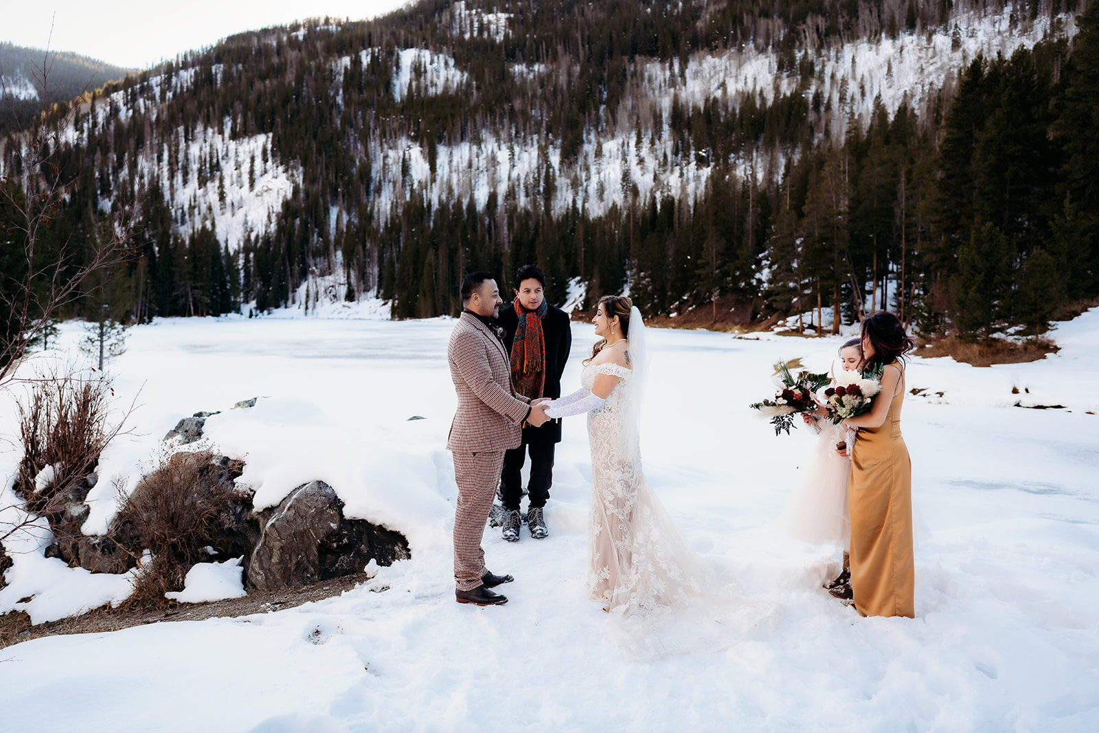 Couple exchanges vows beside a frozen lake surrounded by loved ones and pine trees in the Colorado mountains.
