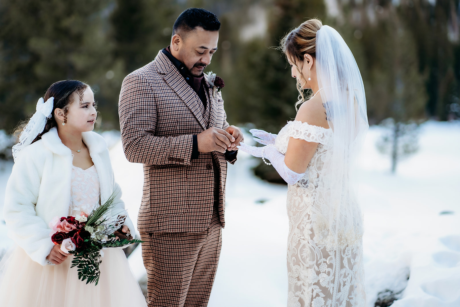 Groom places the ring on the bride’s finger as their daughter watches with joy, standing together in a snowy forest clearing.