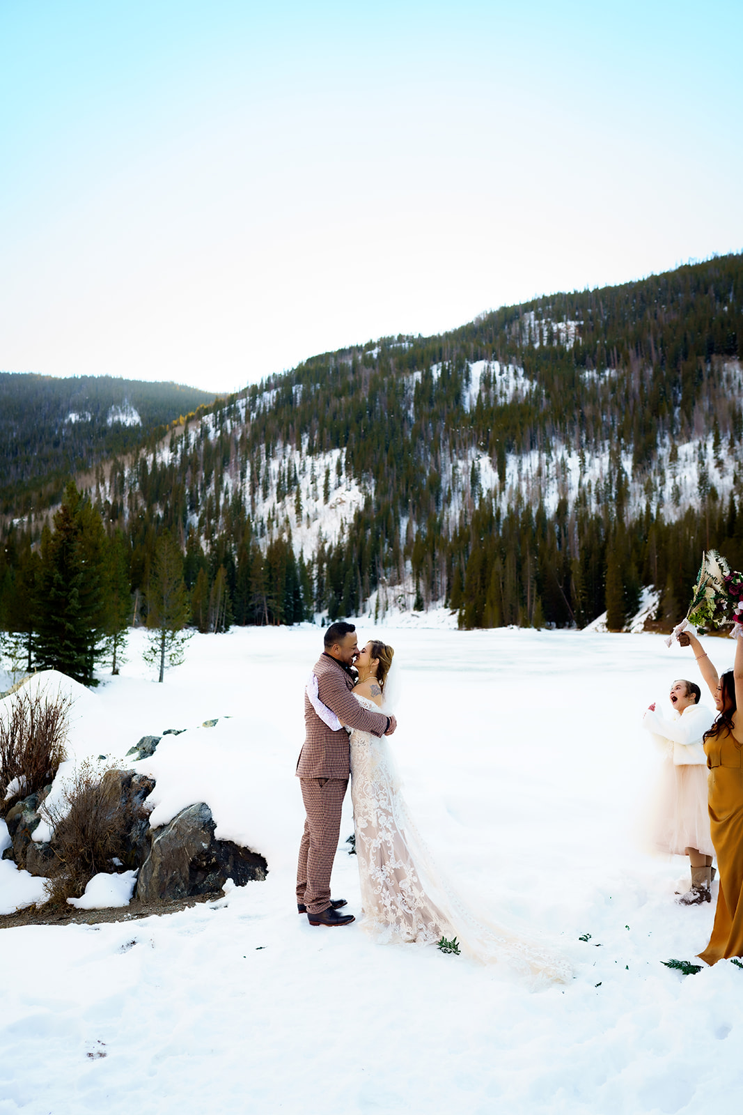 Bride and groom share a kiss as loved ones cheer them on, celebrating their newlywed joy in a snowy Colorado landscape.