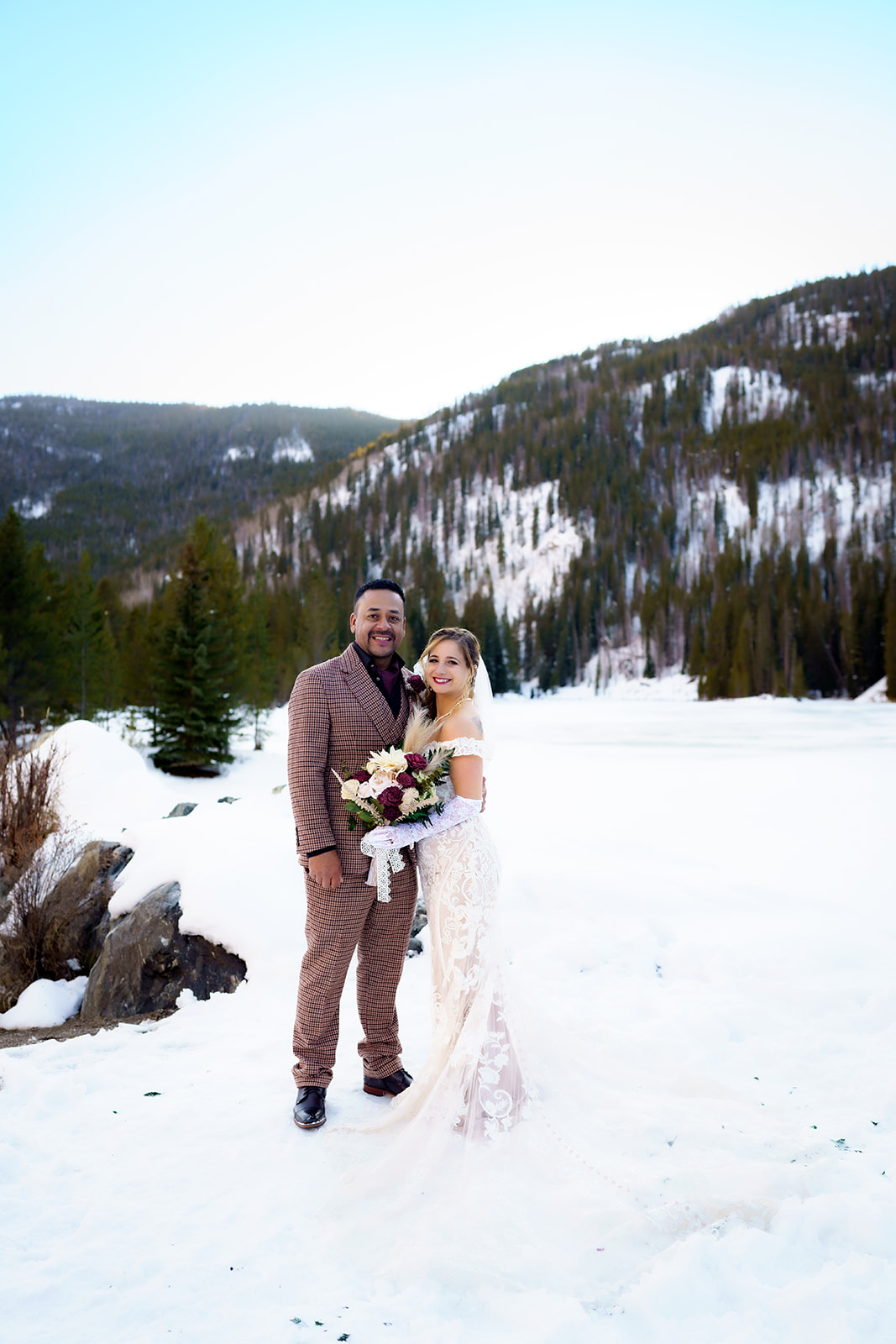 Bride and groom stand joyfully in the snow, with evergreen-covered mountains behind them on their elopement day.