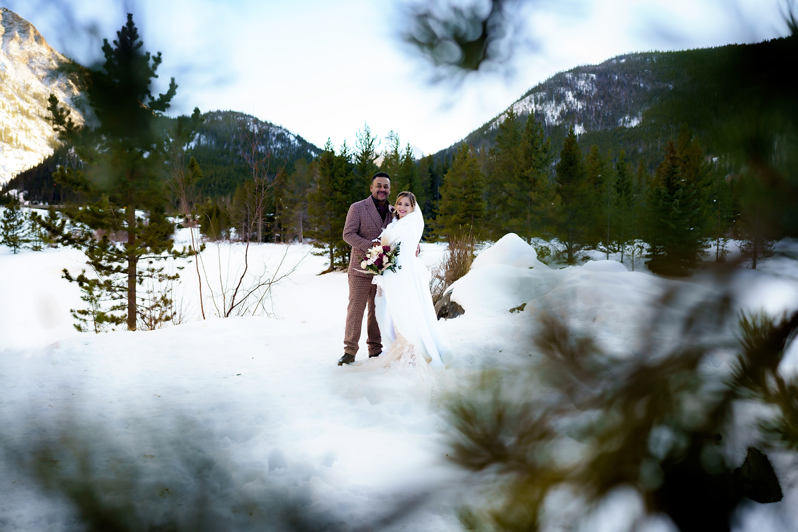 Bride and groom pose through the trees, framed by evergreens and soft snow during their winter elopement in Colorado.