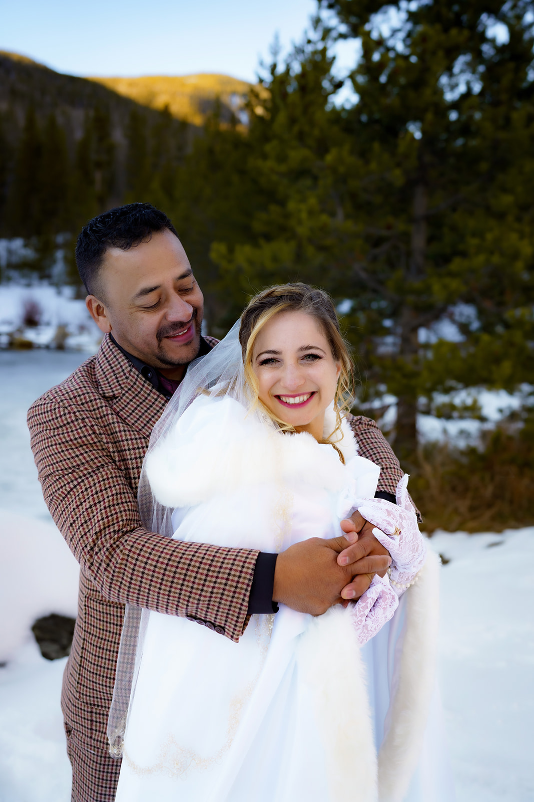 Groom hugs the bride from behind as they smile in a snowy forest clearing during golden hour.