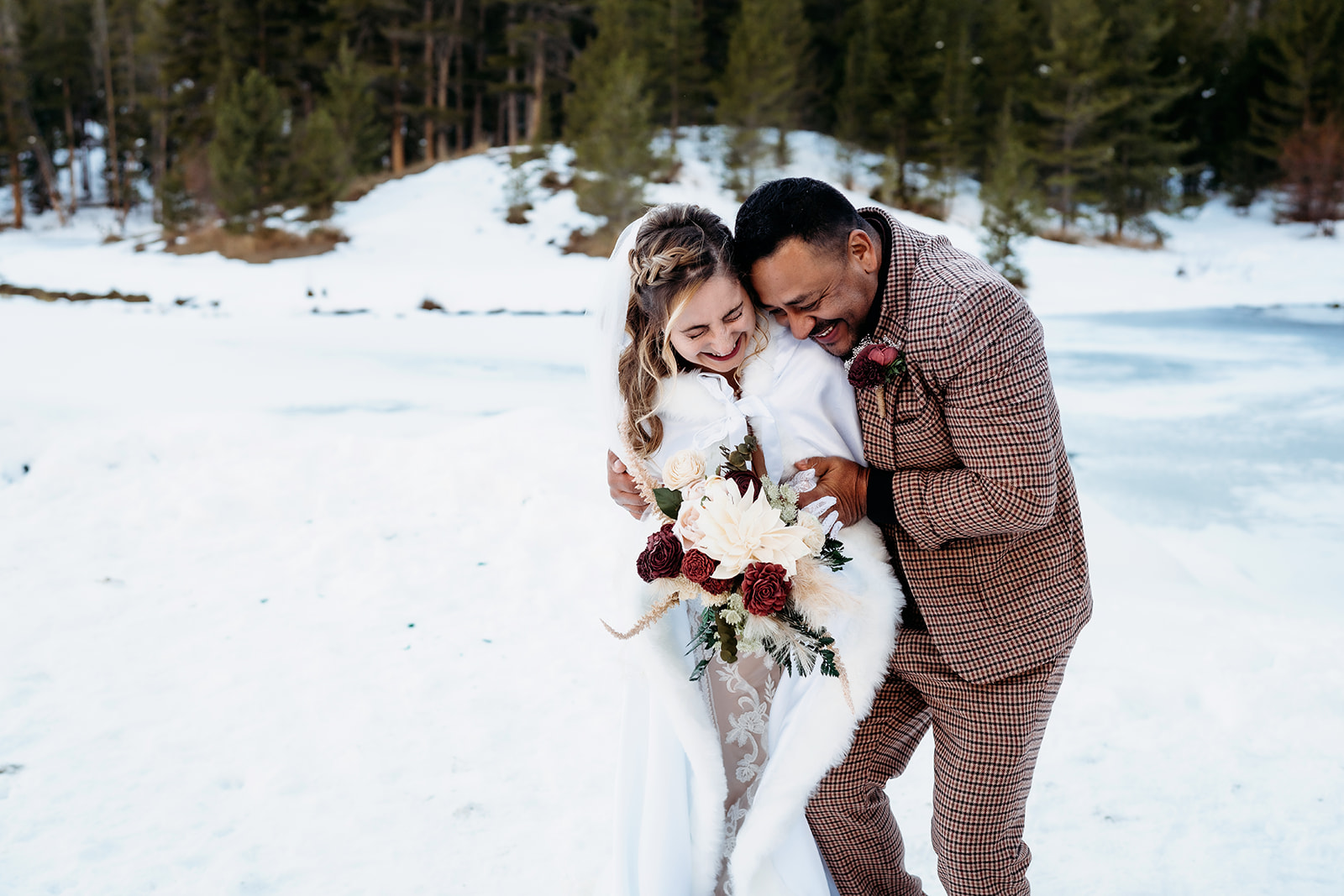 Laughing bride and groom snuggle close in the snow, wrapped in warmth and love.