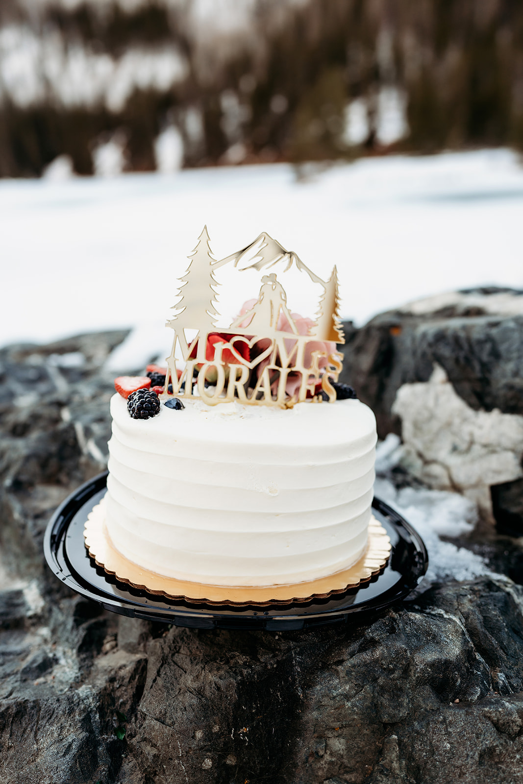 A simple white elopement cake with berries and a custom mountain-themed topper, placed on a rock in the snow.