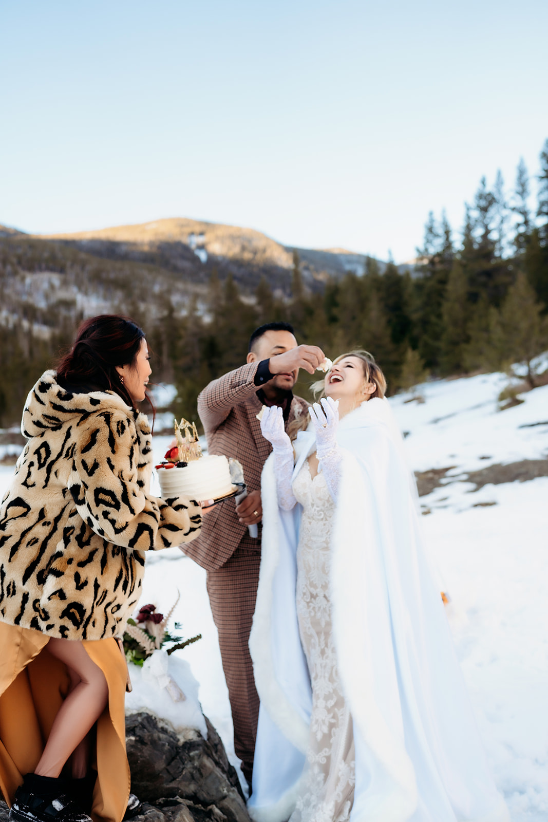Groom feeds the bride a bite of cake while a guest holds the dessert, sharing a playful moment in the winter sunshine.