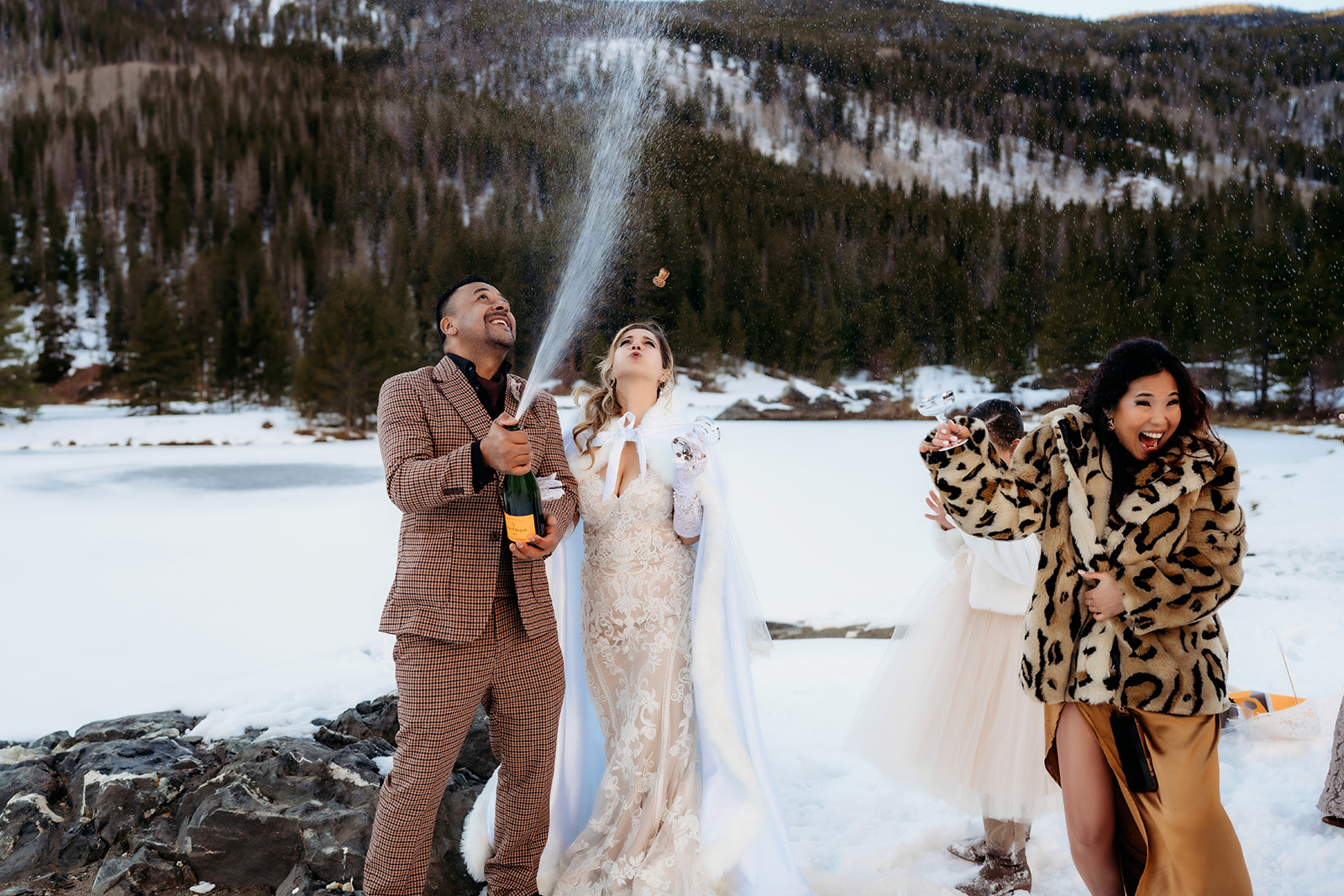 Bride and groom pop a bottle of champagne in celebration, surrounded by laughter and snow in the Colorado mountains.