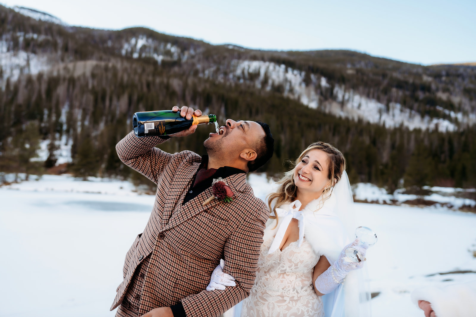 Groom drinks straight from the champagne bottle while the bride laughs beside him during their winter elopement in Colorado.