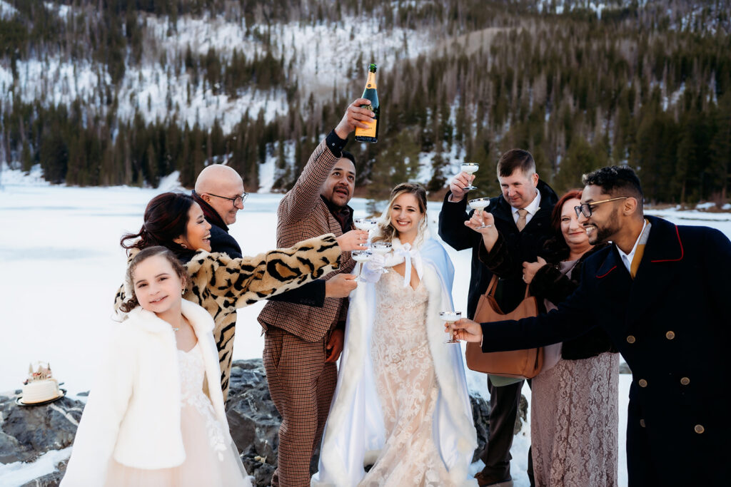 Elopement couple toasts with loved ones beside a frozen Colorado lake, celebrating their winter wedding with champagne.