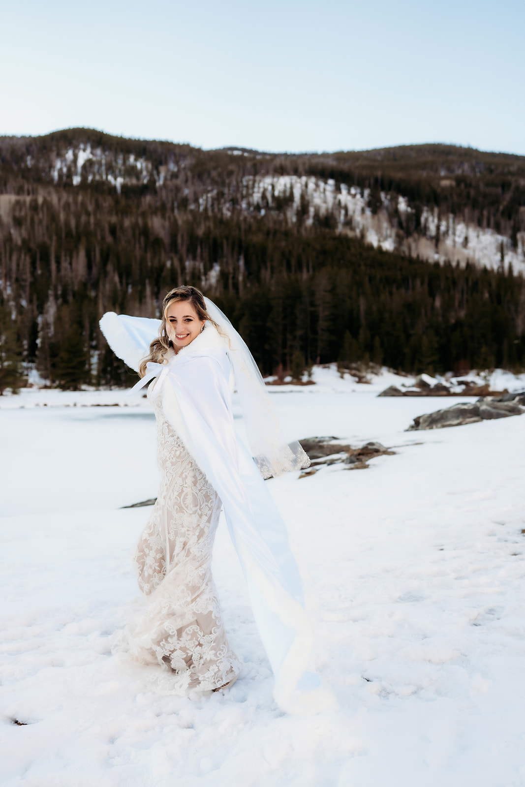 Bride twirls joyfully in the snow in her lace dress and cape, embracing the magic of her winter elopement.