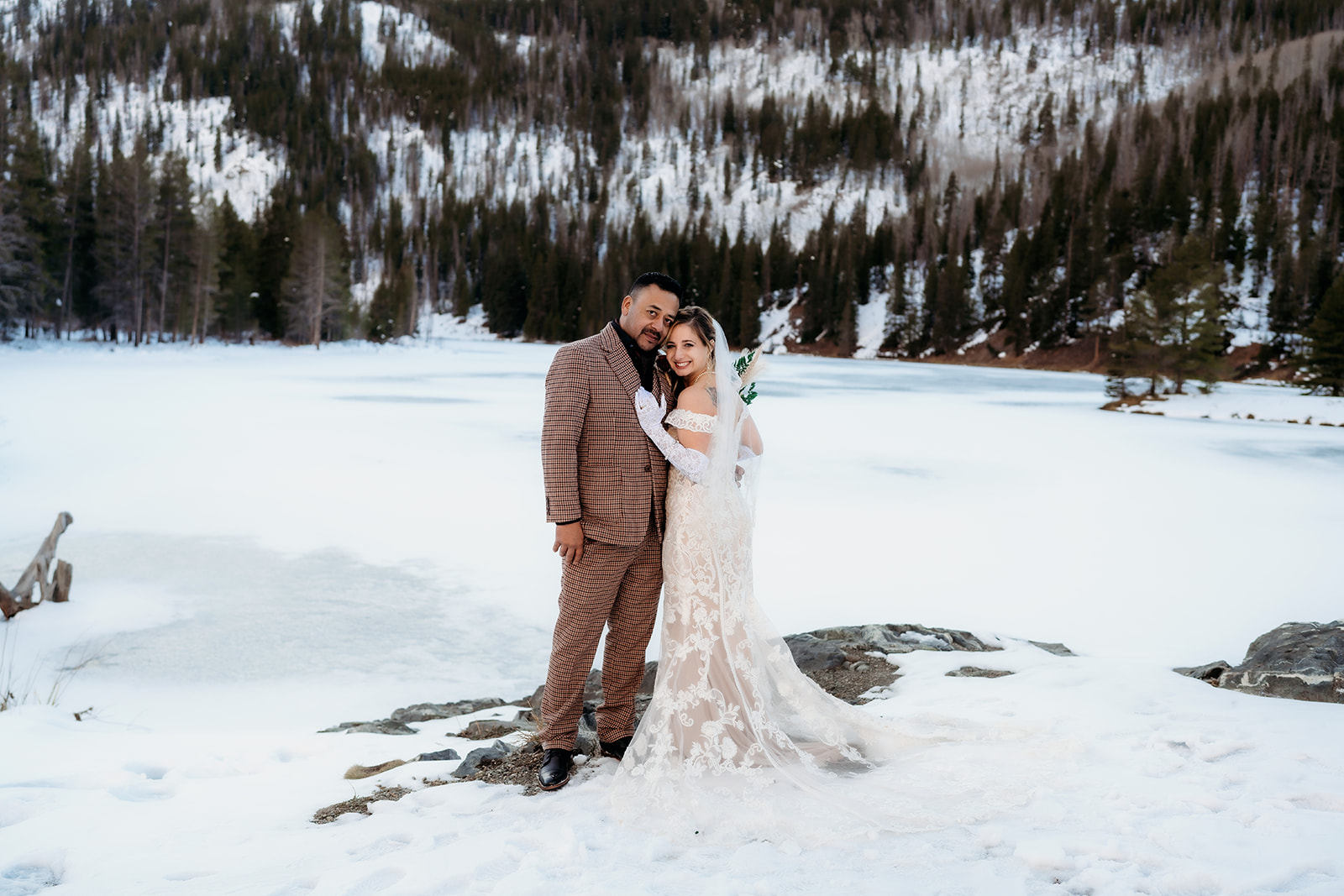 Bride and groom embrace near a frozen lake, mountains rising in the background during their winter elopement in Colorado.