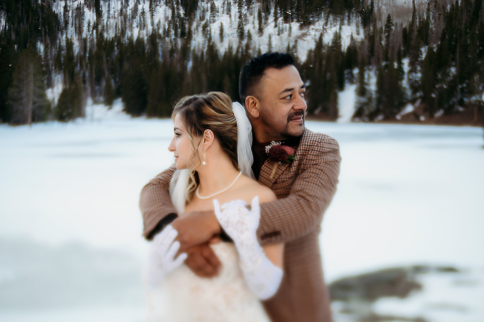 Groom wraps his arms around the bride as they both gaze off into the distance with a snowy lake behind them.